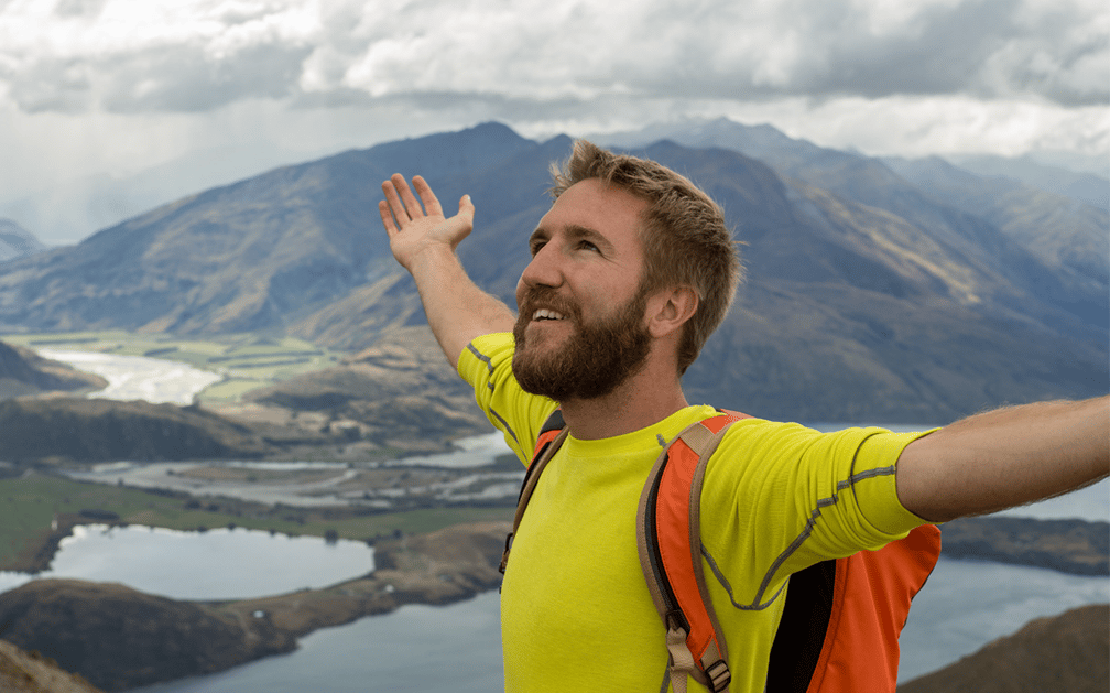 A bearded man with his arms outstretched stands against a mountainous background to signify the relief he’s found from finding the best term life policy possible to protect his family’s financial well-being should he die while he’s still single.