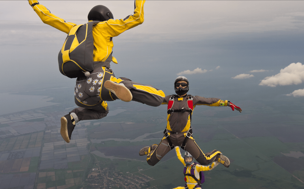 A group of three skydivers with black and yellow jumpsuits and black helmets seems to be floating in the sky above a vast landscape just before they open their parachutes. The picture illustrates daredevil activity and the need for life insurance protection for dangerous hobbies.